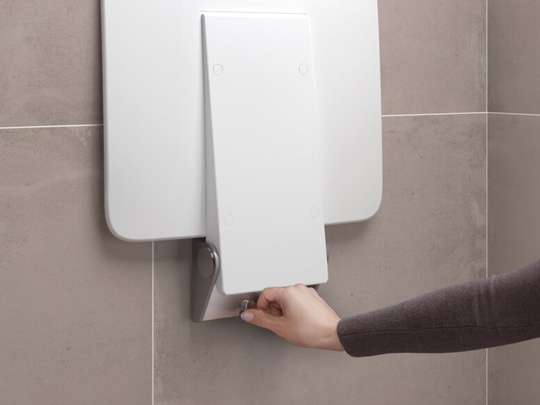 Beige tiled wall with a removable white shower seat that is mounted by one hand